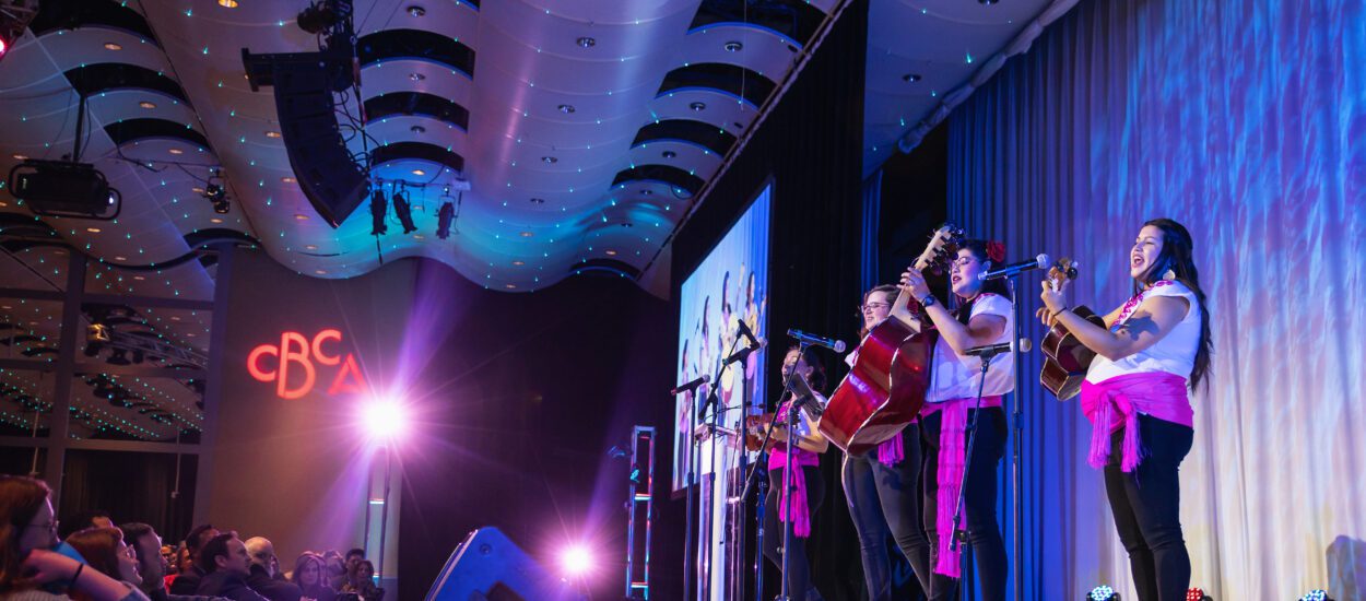 A group of musicians performs on stage with guitars and microphones. They are in a modern, well-lit venue with an audience seated on the left. The letters "CBA" are illuminated on a wall in the background.