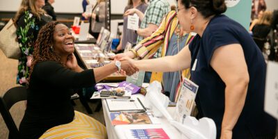 A woman sitting at a table at a job fair enthusiastically shakes hands with a visitor. The table is filled with pamphlets and flyers. In the background, other people are engaged in conversations. The atmosphere is lively and friendly.