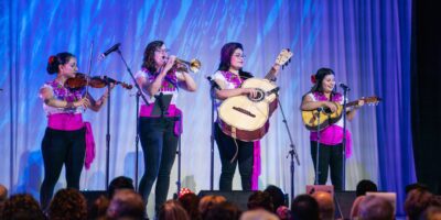 A mariachi band of four women in colorful costumes performs on stage, supported by the Colorado Business Committee for the Arts. One plays the violin, another the trumpet, the third a guitar, and the fourth a vihuela. Blue lighting sets the stage as an attentive audience watches.