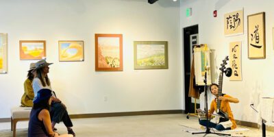 A person sits on the floor of a gallery, playing a stringed instrument while three people watch and listen.