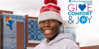 A person wearing a red and gray winter hat and a gray hoodie smiles in front of a hospital building. The text reads "Children's Hospital Colorado Foundation: Give Comfort & Joy" with colorful accents.