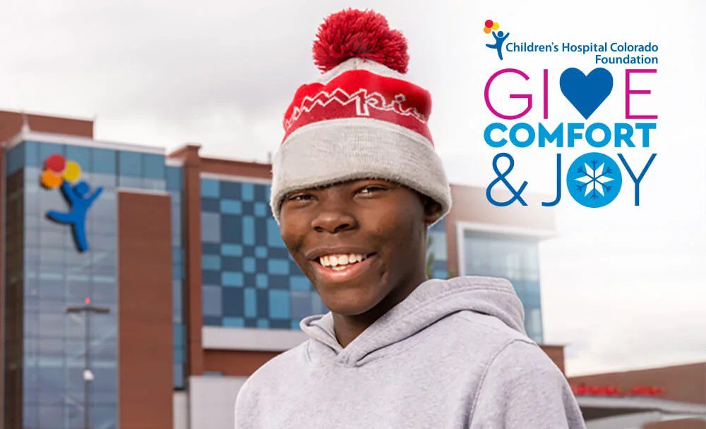 A person wearing a red and gray winter hat and a gray hoodie smiles in front of a hospital building. The text reads "Children's Hospital Colorado Foundation: Give Comfort & Joy" with colorful accents.