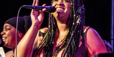 A woman with braided hair sings into a microphone on stage, smiling and gesturing with her hand. Another woman, wearing a headscarf, sings beside her. Both are illuminated by colorful stage lights.