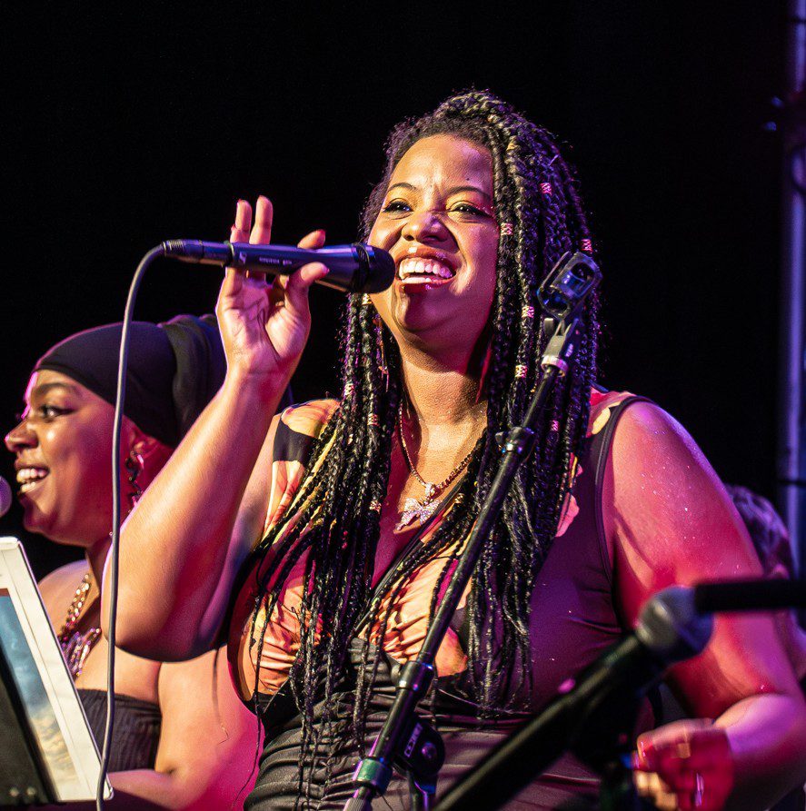 A woman with braided hair sings into a microphone on stage, smiling and gesturing with her hand. Another woman, wearing a headscarf, sings beside her. Both are illuminated by colorful stage lights.