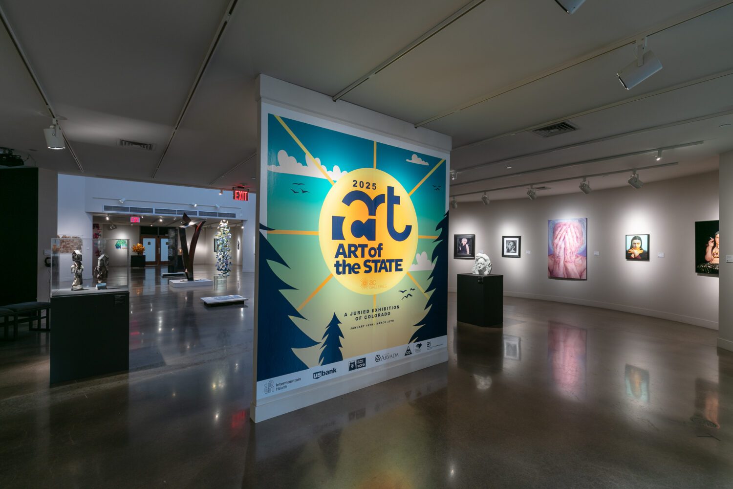 Art gallery interior with various artworks on walls. Prominent poster in center reads "2025 Art of the State: A Juried Exhibition" with a colorful design featuring trees and a sunburst. Dim lighting creates a reflective sheen on the polished floor.