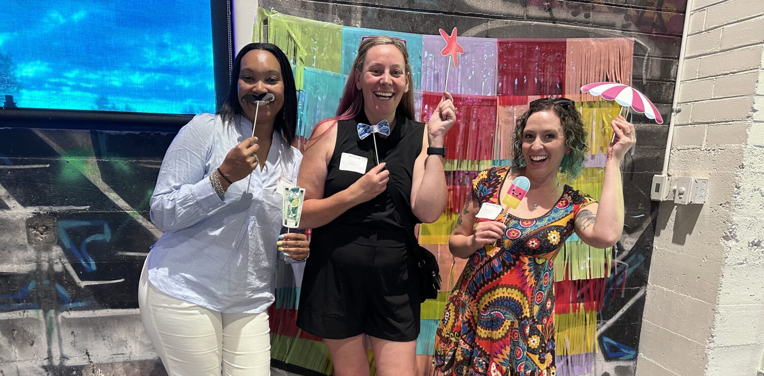 Three women stand smiling in front of a colorful, festive backdrop, holding playful props like a fake mustache, a bow tie, a paper drink, and a small umbrella, creating a cheerful and fun atmosphere indoors.