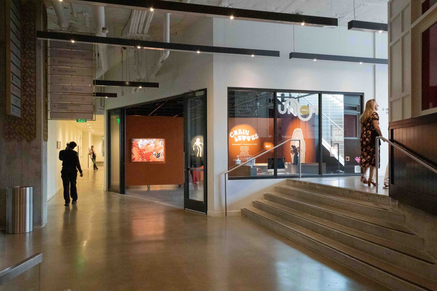 A modern museum interior with polished concrete floors and angled walls. Exhibits include "CORAL REEFERS" and a colorful art display. A woman stands on steps, and a person walks through the hallway. Ceiling lights illuminate the space.