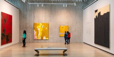 A modern art gallery features large abstract paintings with red, yellow, and black tones. Three people observe the art on the walls, while a fourth person stands near a wooden bench in the center of the spacious room.