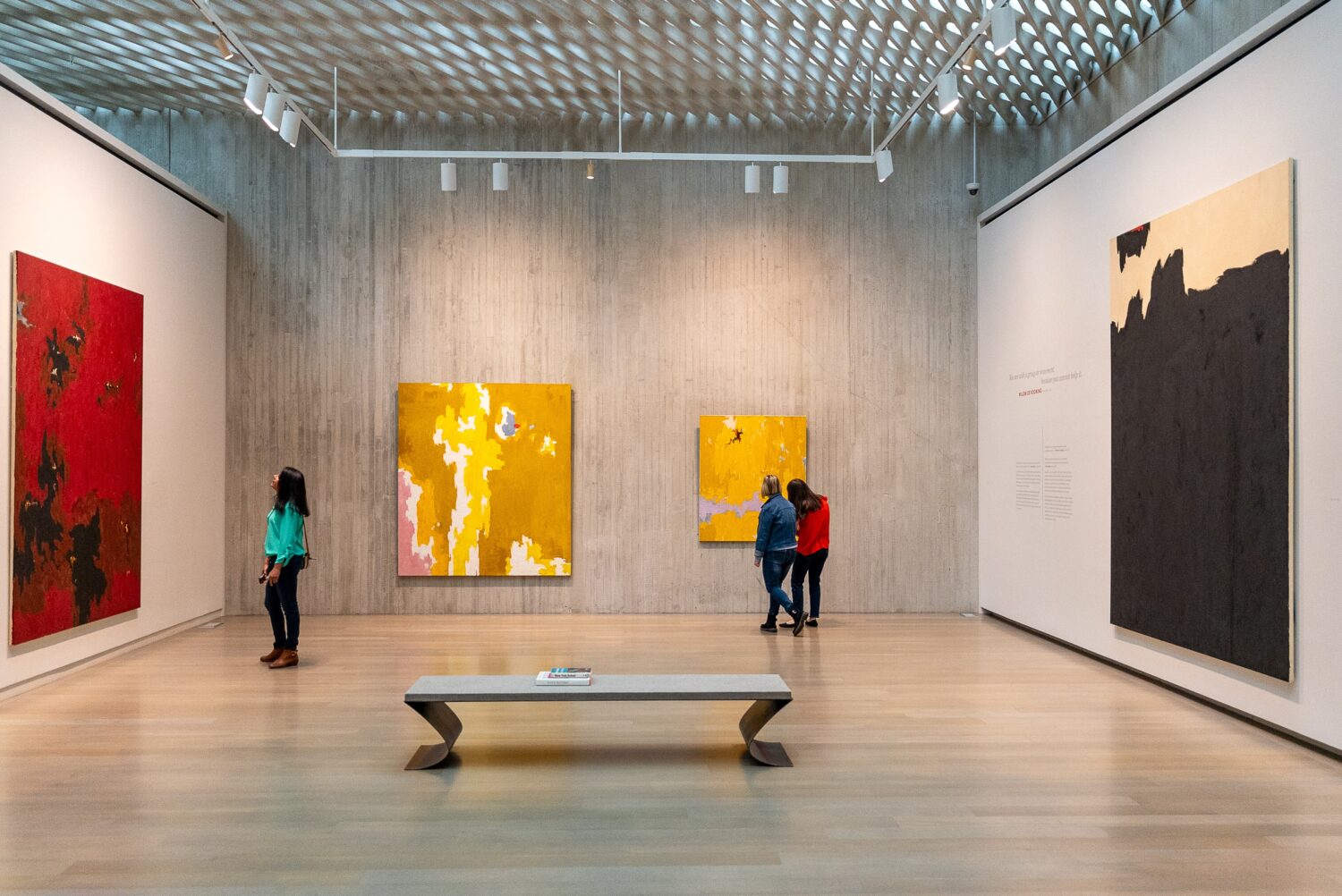 A modern art gallery features large abstract paintings with red, yellow, and black tones. Three people observe the art on the walls, while a fourth person stands near a wooden bench in the center of the spacious room.