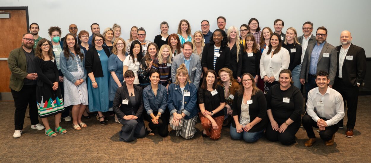 A large group of people, dressed in business casual attire, pose together and smile for a group photo in a conference room with a plain white wall and brown carpet.