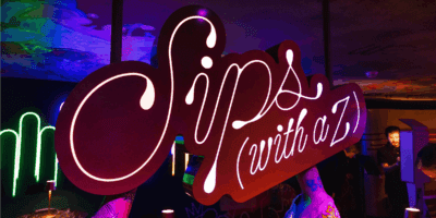 A large, illuminated sign with the words “Sips (with a Z)” in cursive lettering is displayed in a vibrant, colorful indoor setting with neon lights and people in the background.