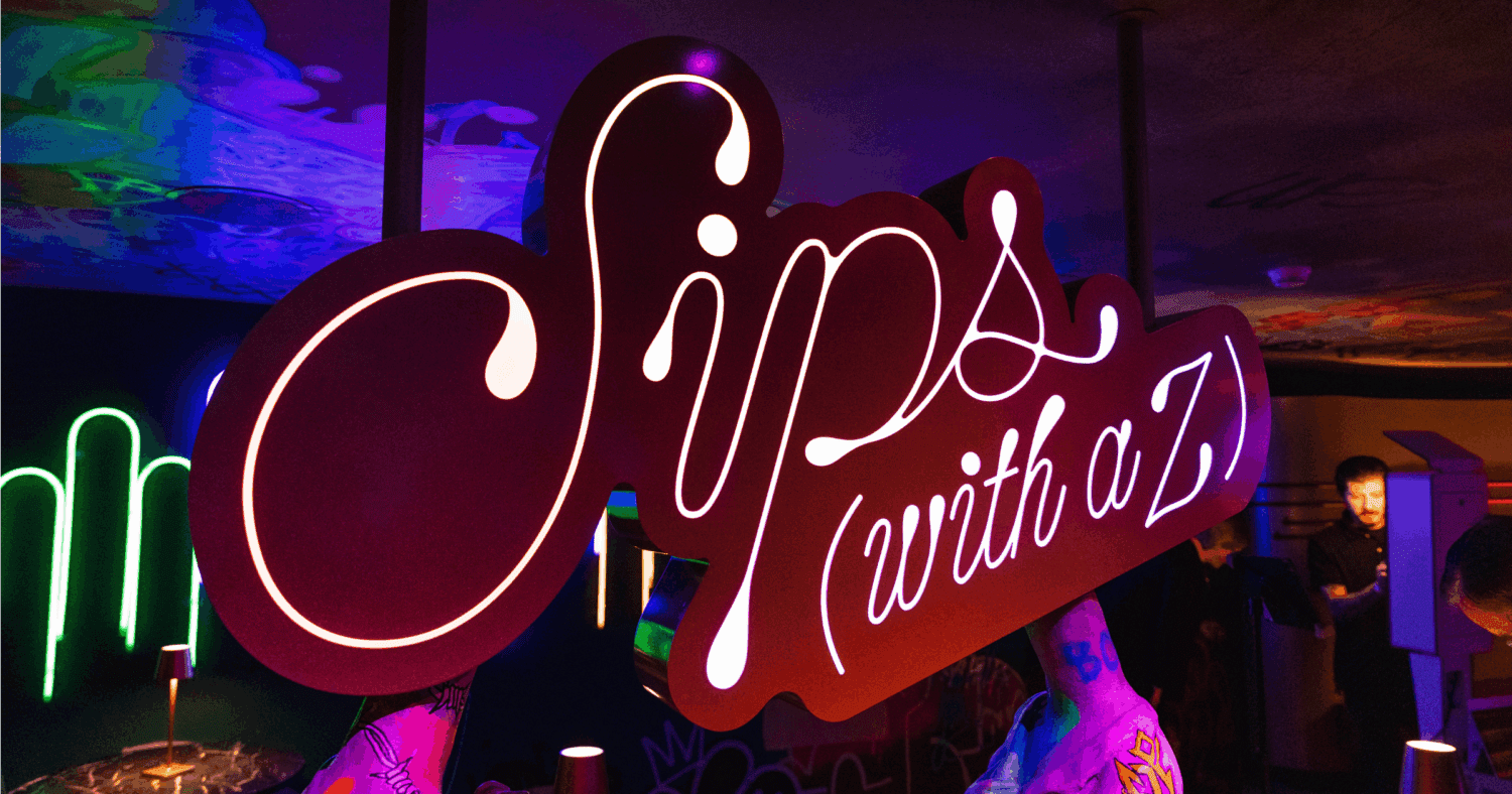 A large, illuminated sign with the words “Sips (with a Z)” in cursive lettering is displayed in a vibrant, colorful indoor setting with neon lights and people in the background.