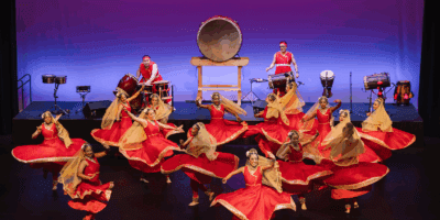 A group of dancers in red and gold costumes perform energetically on stage, spinning with raised arms, while musicians in matching attire play large drums and percussion instruments against a purple-lit background.