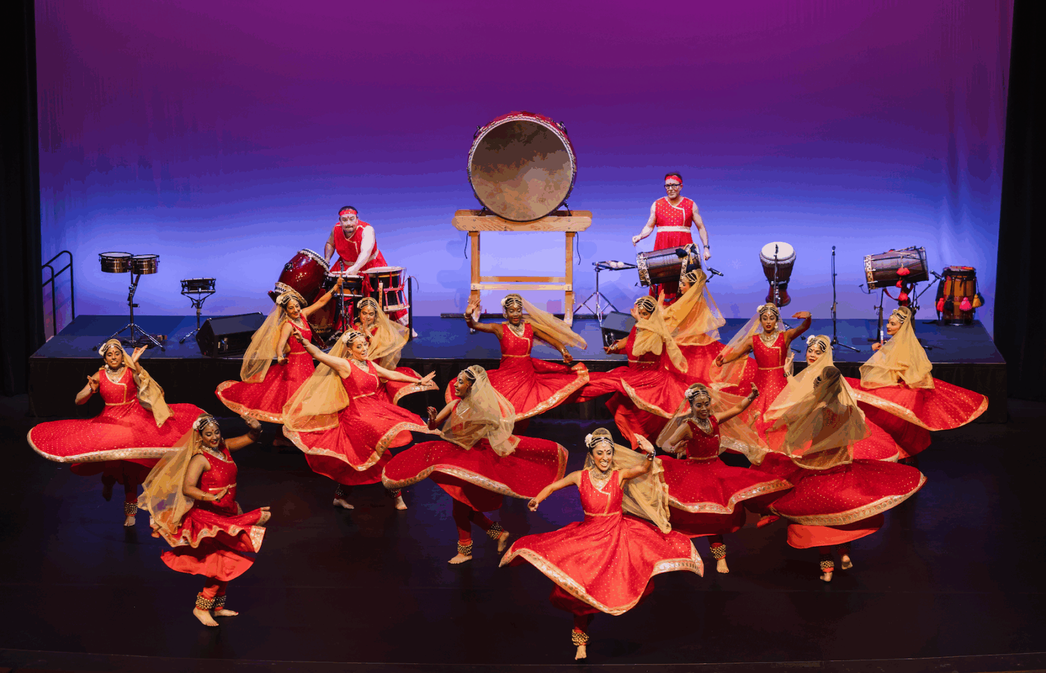 A group of dancers in red and gold costumes perform energetically on stage, spinning with raised arms, while musicians in matching attire play large drums and percussion instruments against a purple-lit background.