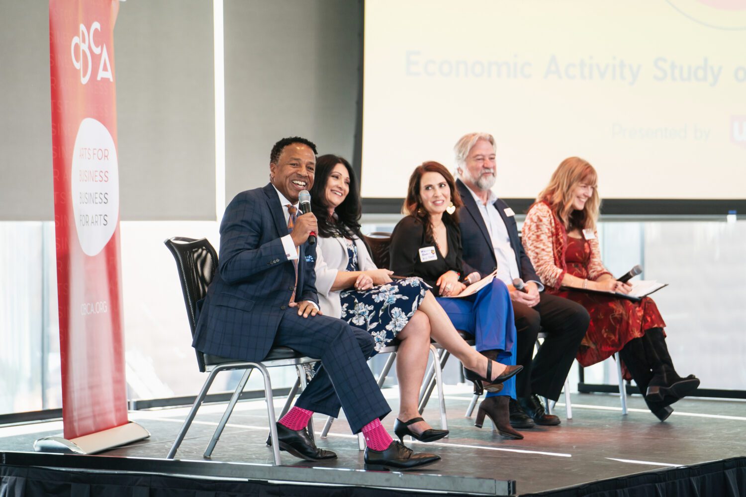 Five panelists sit on stage, smiling and holding microphones and papers during a business event. A large presentation screen and a red banner are visible in the background. The setting is bright and professional.