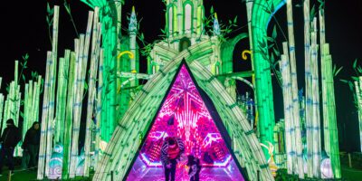 Two people stand inside a brightly lit, triangular tunnel with vibrant pink and purple lights, surrounded by tall, glowing green bamboo structures at night.
