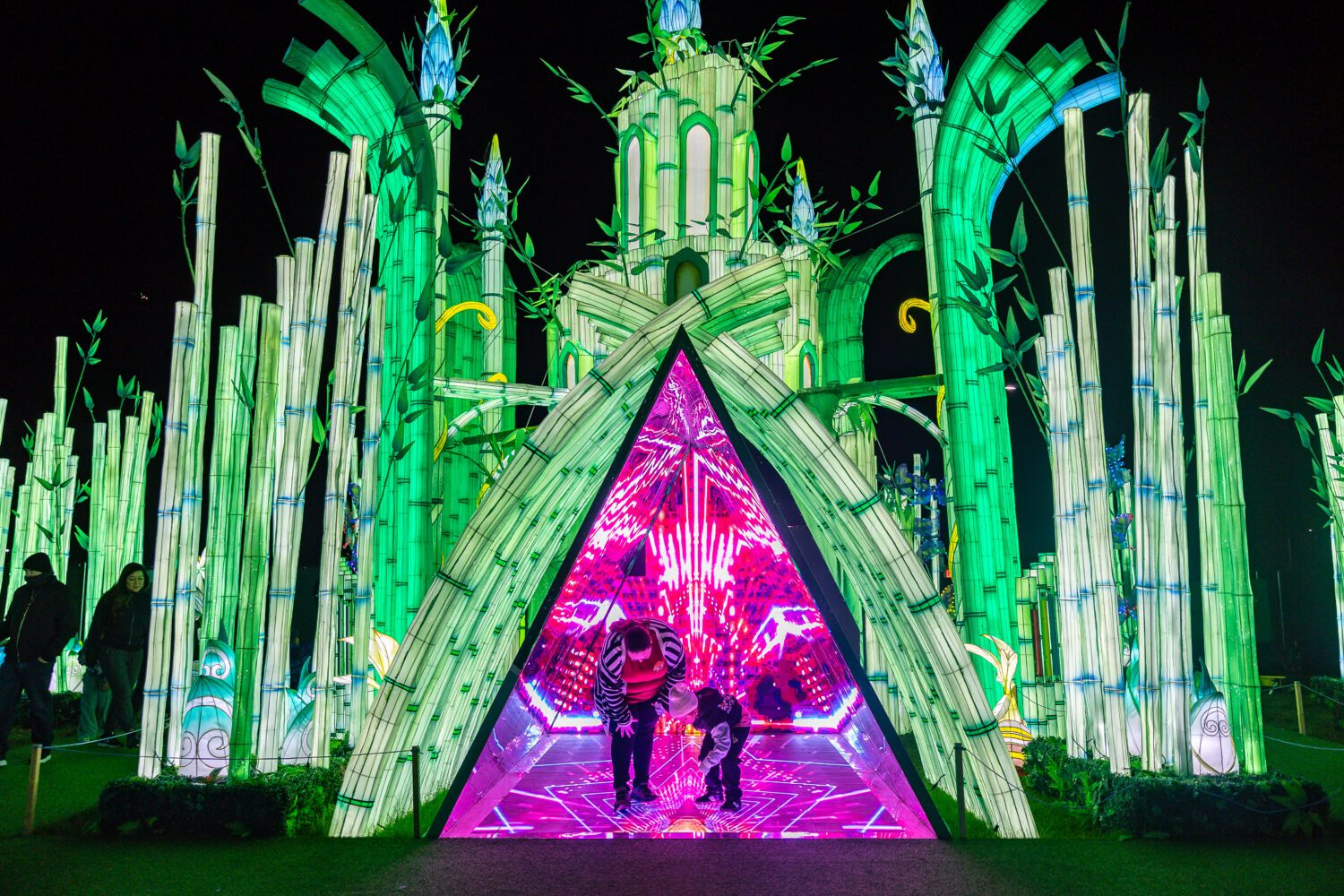 Two people stand inside a brightly lit, triangular tunnel with vibrant pink and purple lights, surrounded by tall, glowing green bamboo structures at night.