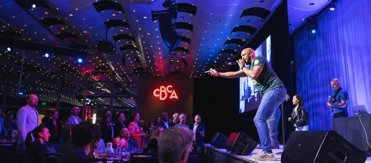 A performer energetically sings on stage at an indoor event, engaging with a standing audience. The venue has modern lighting, a high ceiling, and a glowing “CBA” sign in the background. People watch from tables and the floor.