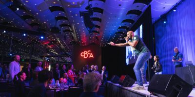 A performer energetically sings on stage at an indoor event, engaging with a standing audience. The venue has modern lighting, a high ceiling, and a glowing “CBA” sign in the background. People watch from tables and the floor.