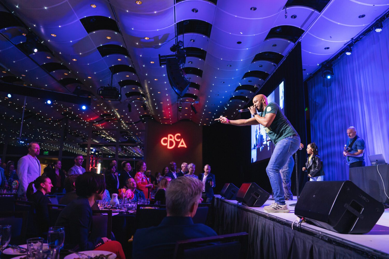 A performer energetically sings on stage at an indoor event, engaging with a standing audience. The venue has modern lighting, a high ceiling, and a glowing “CBA” sign in the background. People watch from tables and the floor.