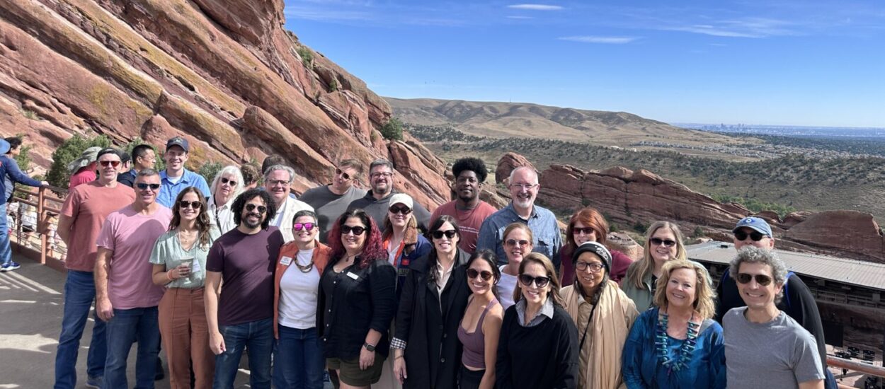 A group of about 25 people posing and smiling outdoors at Red Rocks Amphitheatre, with large red rock formations and distant hills under a blue sky in the background.