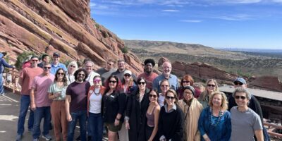 A group of about 25 people posing and smiling outdoors at Red Rocks Amphitheatre, with large red rock formations and distant hills under a blue sky in the background.