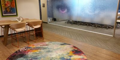 A modern office lounge with a colorful round rug, a wooden table with four chairs, a framed artwork on the wall, and a glass meeting room with large eyes printed on the frosted glass.