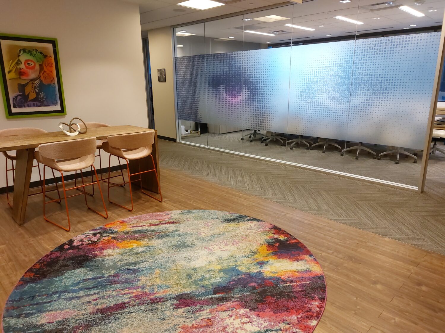 A modern office lounge with a colorful round rug, a wooden table with four chairs, a framed artwork on the wall, and a glass meeting room with large eyes printed on the frosted glass.