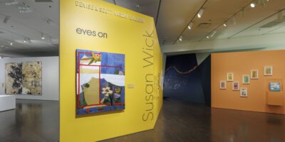 A brightly lit art gallery features a large yellow wall displaying a colorful abstract painting and the text “eyes on Susan Wick.” Other artworks hang on adjacent yellow and white walls in the spacious room.
