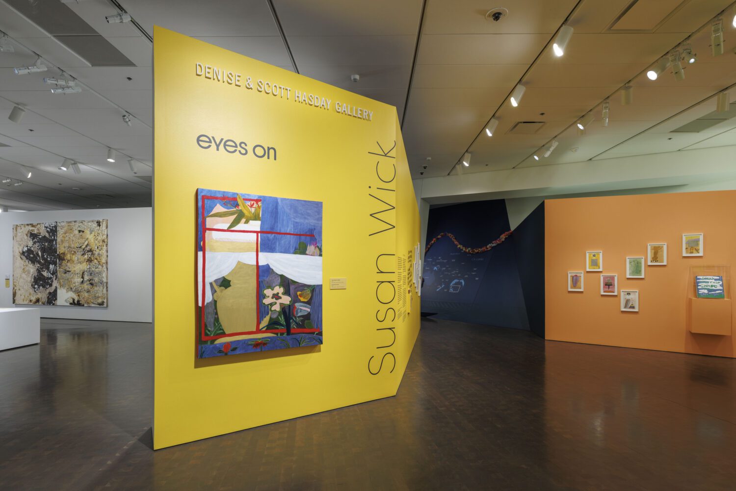 A brightly lit art gallery features a large yellow wall displaying a colorful abstract painting and the text “eyes on Susan Wick.” Other artworks hang on adjacent yellow and white walls in the spacious room.