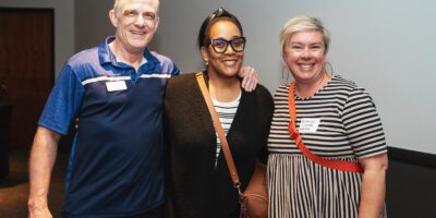 Three people smiling and posing together indoors; a man in a blue polo shirt stands beside two women, one wearing glasses and black clothing, the other in a striped dress with a red bag.