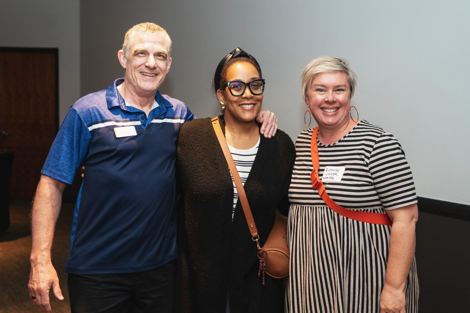 Three people smiling and posing together indoors; a man in a blue polo shirt stands beside two women, one wearing glasses and black clothing, the other in a striped dress with a red bag.
