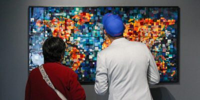 Two people stand closely together, facing a colorful, abstract mosaic artwork made of small squares hanging on a gray wall. One wears a red sweater, the other a white jacket and blue cap.
