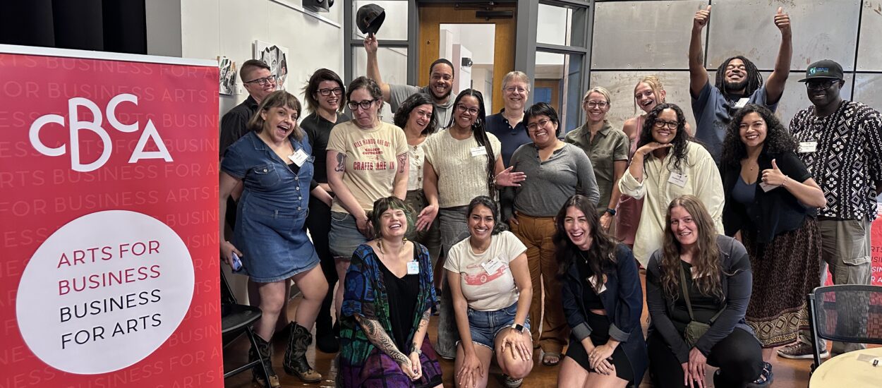 A diverse group of people pose and smile together indoors next to a pink CBCA banner that reads "Arts for Business, Business for Arts." Several people give thumbs up and appear happy and energetic.