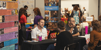 People sit at tables at a busy indoor event or fair, talking and sharing information. Colorful posters and banners with text and images are displayed on the tables and walls. Several people stand and walk in the background.