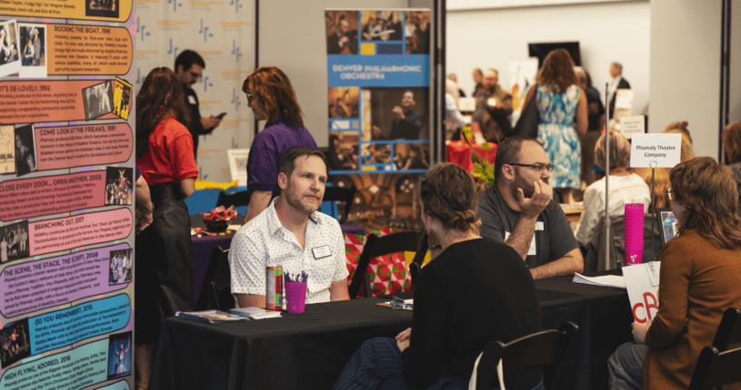 People sit at tables at a busy indoor event or fair, talking and sharing information. Colorful posters and banners with text and images are displayed on the tables and walls. Several people stand and walk in the background.