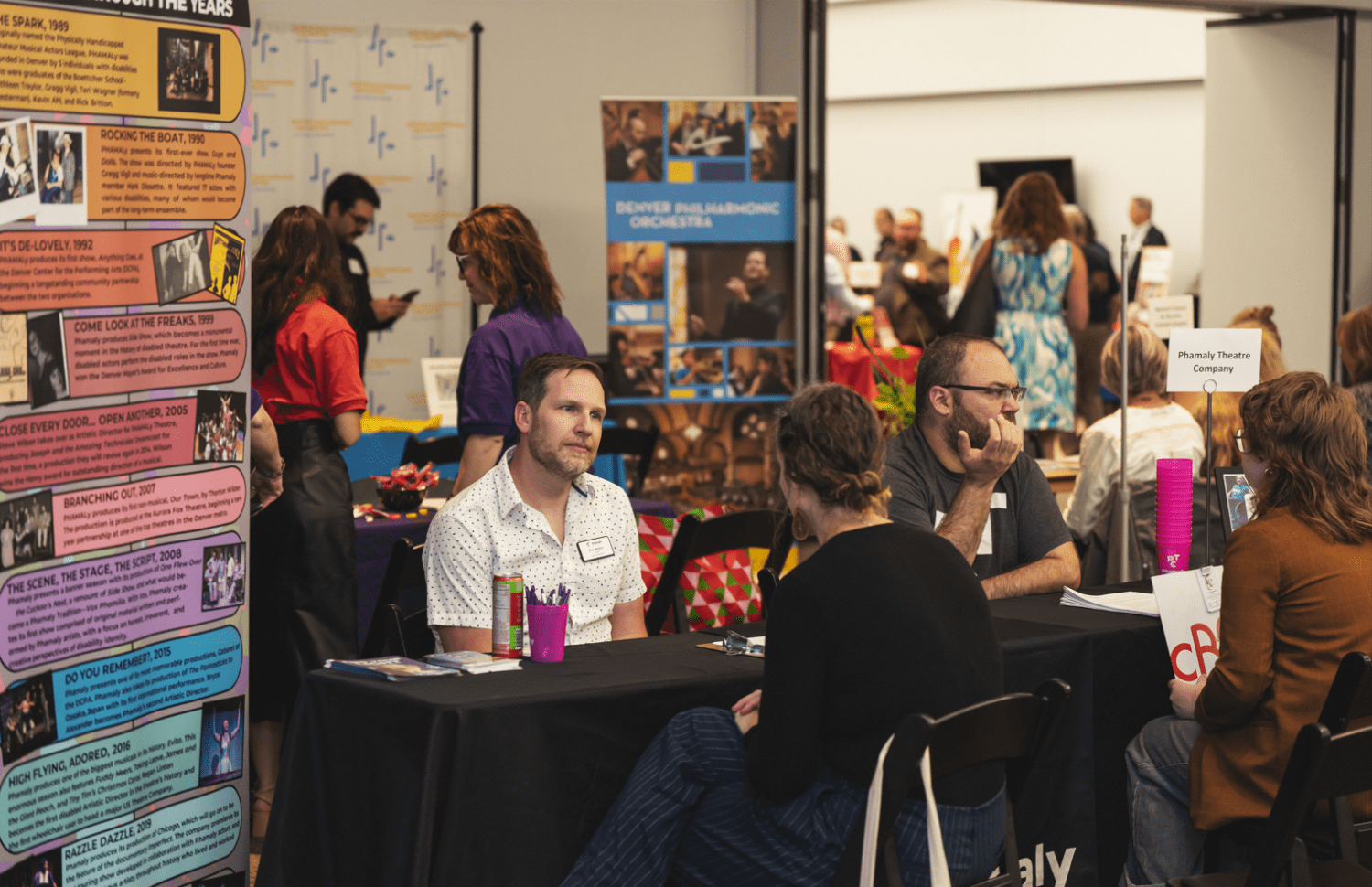 People sit at tables at a busy indoor event or fair, talking and sharing information. Colorful posters and banners with text and images are displayed on the tables and walls. Several people stand and walk in the background.