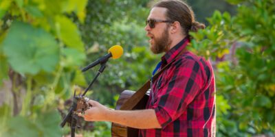 A man with a beard and sunglasses, wearing a red plaid shirt, plays an acoustic guitar and sings into a microphone with a yellow foam cover amid lush green outdoor surroundings.