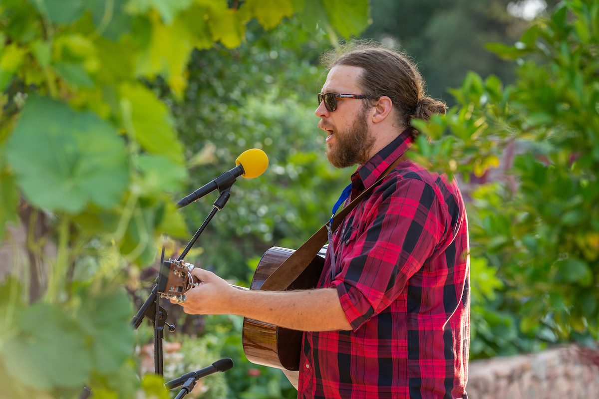 A man with a beard and sunglasses, wearing a red plaid shirt, plays an acoustic guitar and sings into a microphone with a yellow foam cover amid lush green outdoor surroundings.
