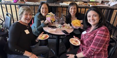 Four women sit around a small table at a restaurant, smiling at the camera. They have plates of food and glasses of wine, with two women holding flower bouquets. Name tags are visible on their shirts.