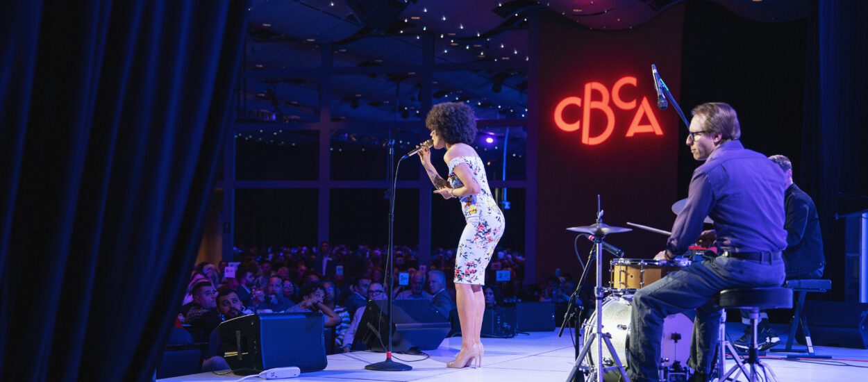 A woman sings on stage with a band; a drummer and keyboardist play beside her. The stage is lit with blue and purple lights, and a large crowd watches. The illuminated letters "CBCA" are displayed in the background.
