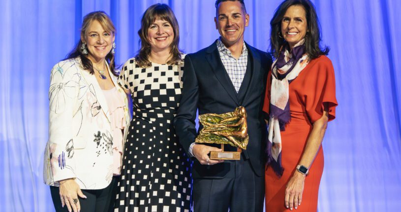 Four people stand together, smiling in front of a blue curtain. One man, holding a gold award from the Colorado Business Committee for the Arts, is flanked by three women dressed in business attire.