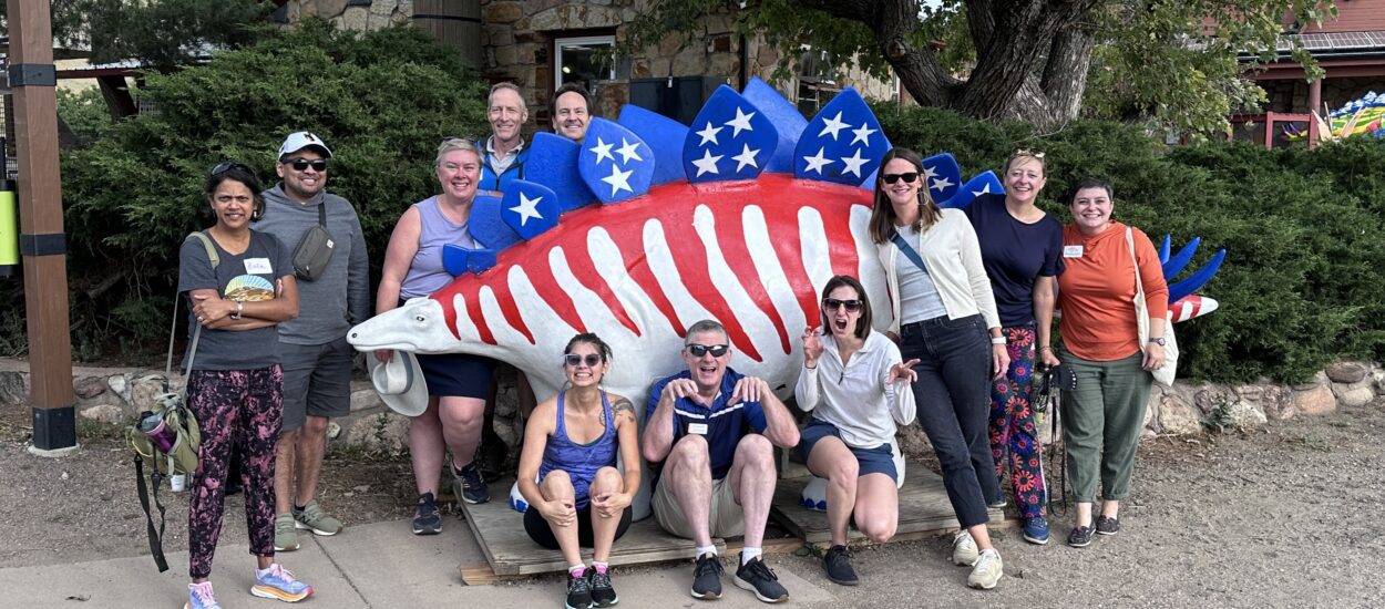 A group of thirteen smiling people pose in front of a statue of a stegosaurus painted with red stripes and blue plates decorated with white stars, resembling the American flag. Trees and a stone building are in the background.