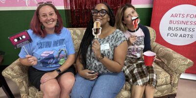 Three women sit on a patterned couch, smiling and holding playful props including a microphone, lips, and a "1 UP" sign. Behind them are colorful pink, green, and red decorations and a "Business for Arts" banner.