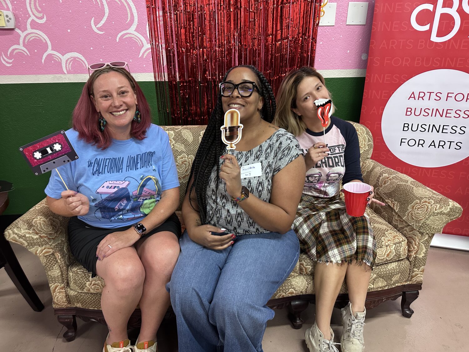 Three women sit on a patterned couch, smiling and holding playful props including a microphone, lips, and a "1 UP" sign. Behind them are colorful pink, green, and red decorations and a "Business for Arts" banner.
