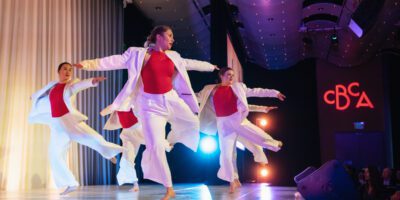 Four dancers in white outfits and red tops perform a synchronized routine on a brightly lit stage, with arms extended gracefully. The background features stage lighting and the letters "CBCA" illuminated in red.