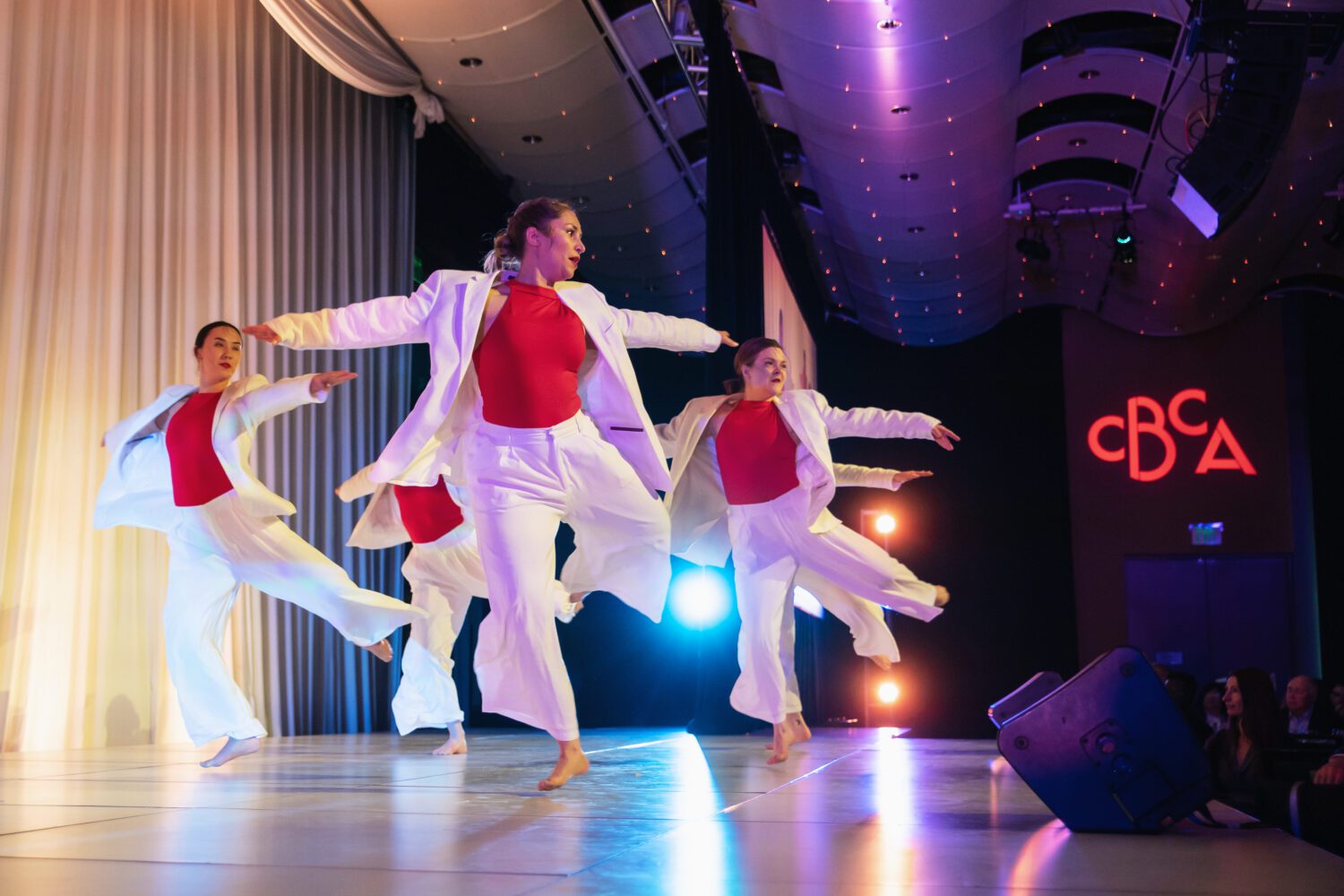 Four dancers in white outfits and red tops perform a synchronized routine on a brightly lit stage, with arms extended gracefully. The background features stage lighting and the letters "CBCA" illuminated in red.