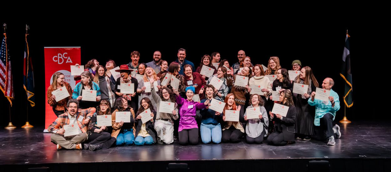 A large group of people pose on a stage, smiling and holding certificates. Flags and a red banner are in the background, suggesting a celebratory or graduation event.