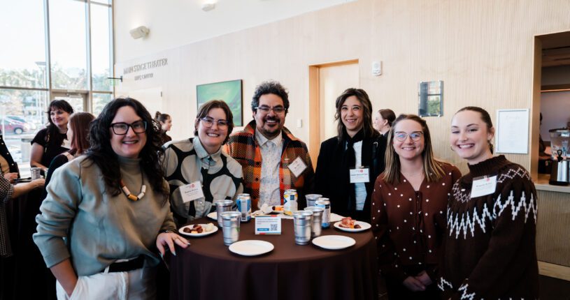 Six people with name tags smile around a tall round table with food and drinks in a bright, modern room adorned with art. They appear to be attending a Colorado Business Committee for the Arts event in smart business casual attire.