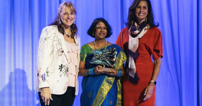 Three women pose together in front of blue curtains. The woman in the center, dressed in a blue and gold sari, holds a Colorado Business Committee for the Arts trophy. The women on either side wear a white jacket and red dress, smiling warmly.